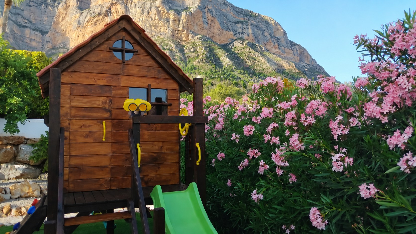 Casita infantil de madera con tobogán verde, flores rosas y montañas